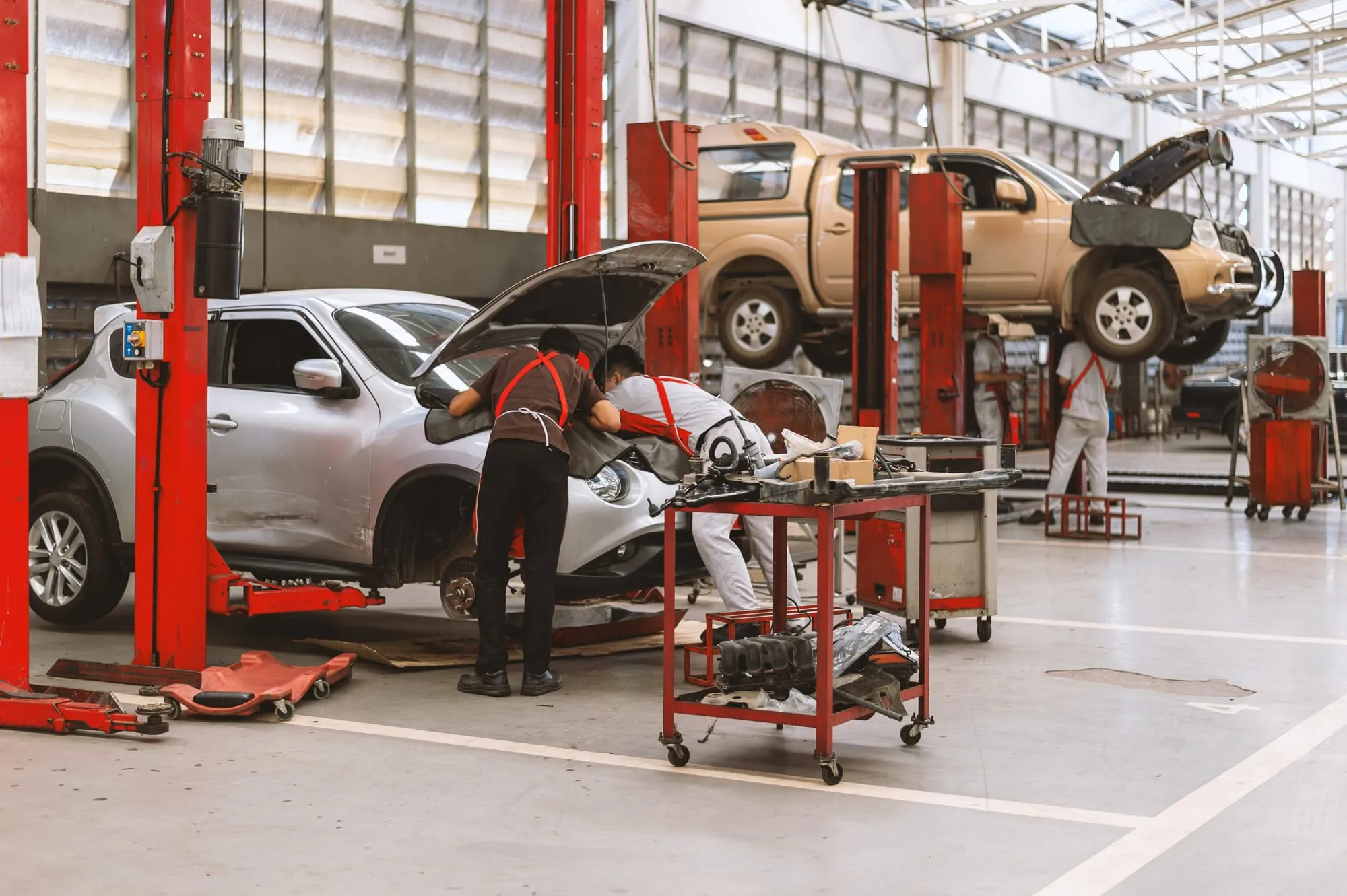 Deux mécaniciens interviennent sous le capot d’un véhicule dans un atelier, avec un pick-up sur un pont en arrière-plan.